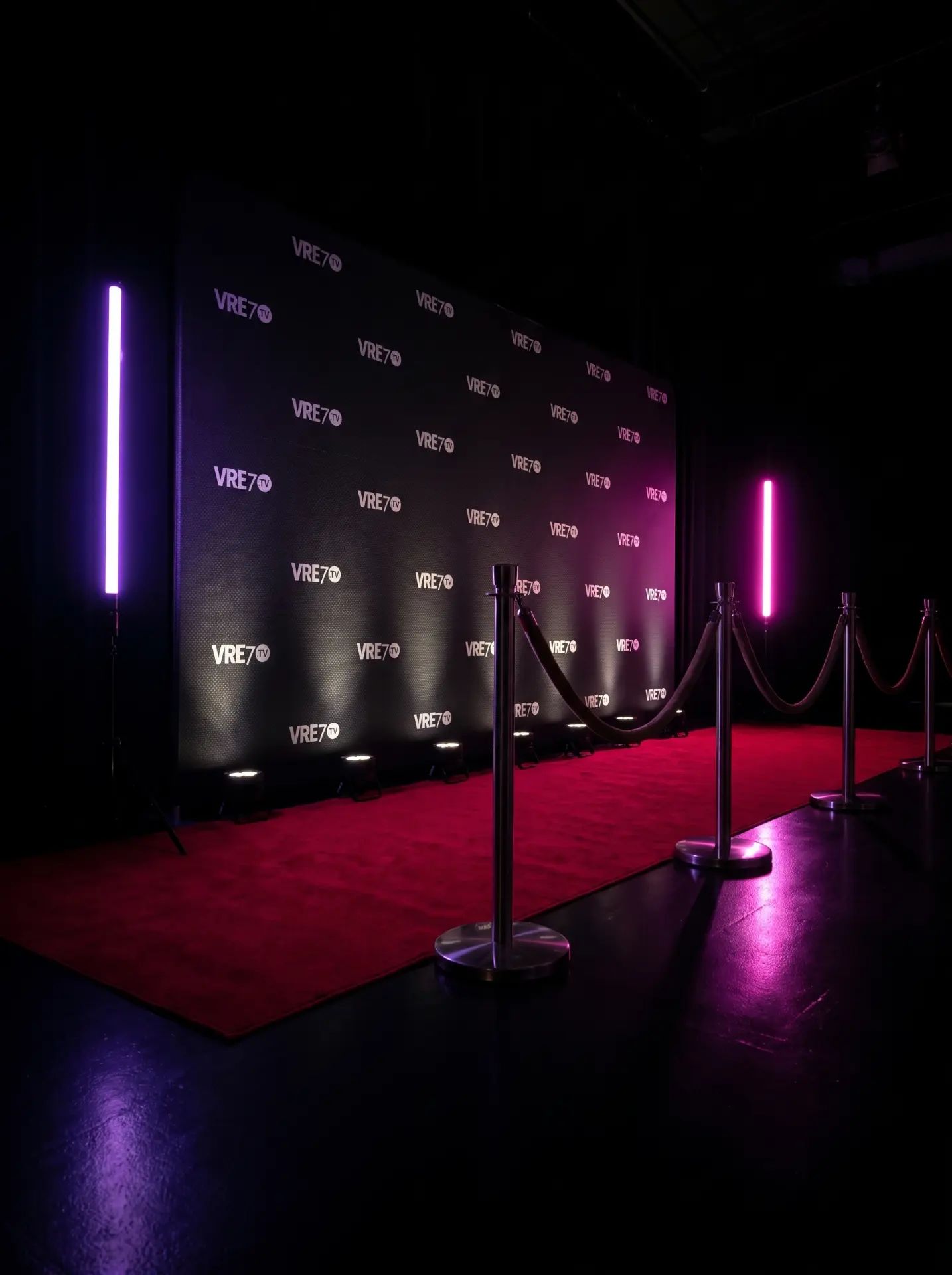 A red carpet with velvet ropes leading to the VRE7 TV step-and-repeat wall, lit by purple and pink neon tubes